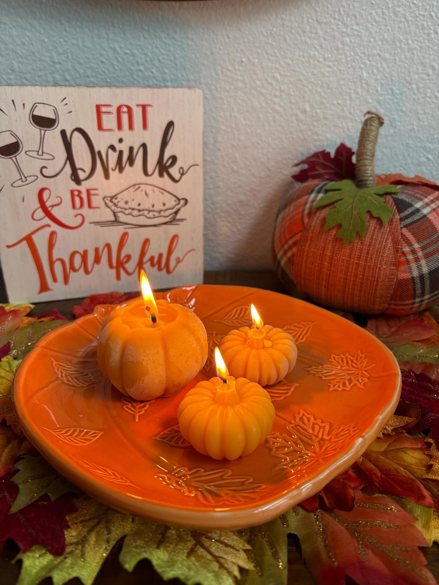 Decorative setting with lit beeswax candles shaped like pumpkins on an orange plate, surrounded by fall leaves and a sign that reads 'Eat Drink & Be Thankful'.