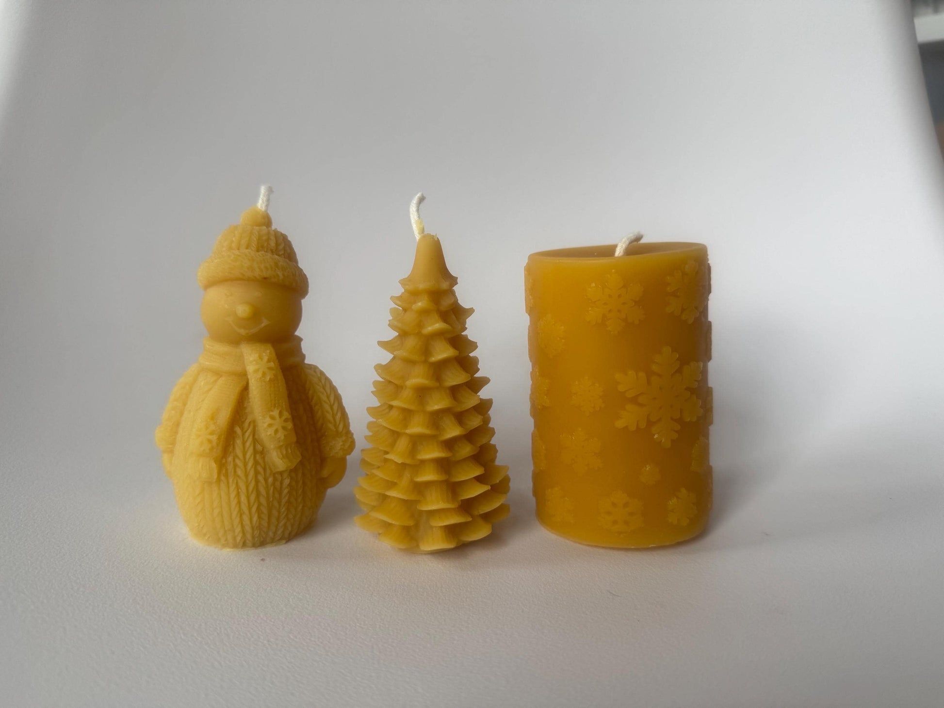 Three beeswax candles shaped like a snowman, tree, and cylindrical design with snowflakes on a white background.